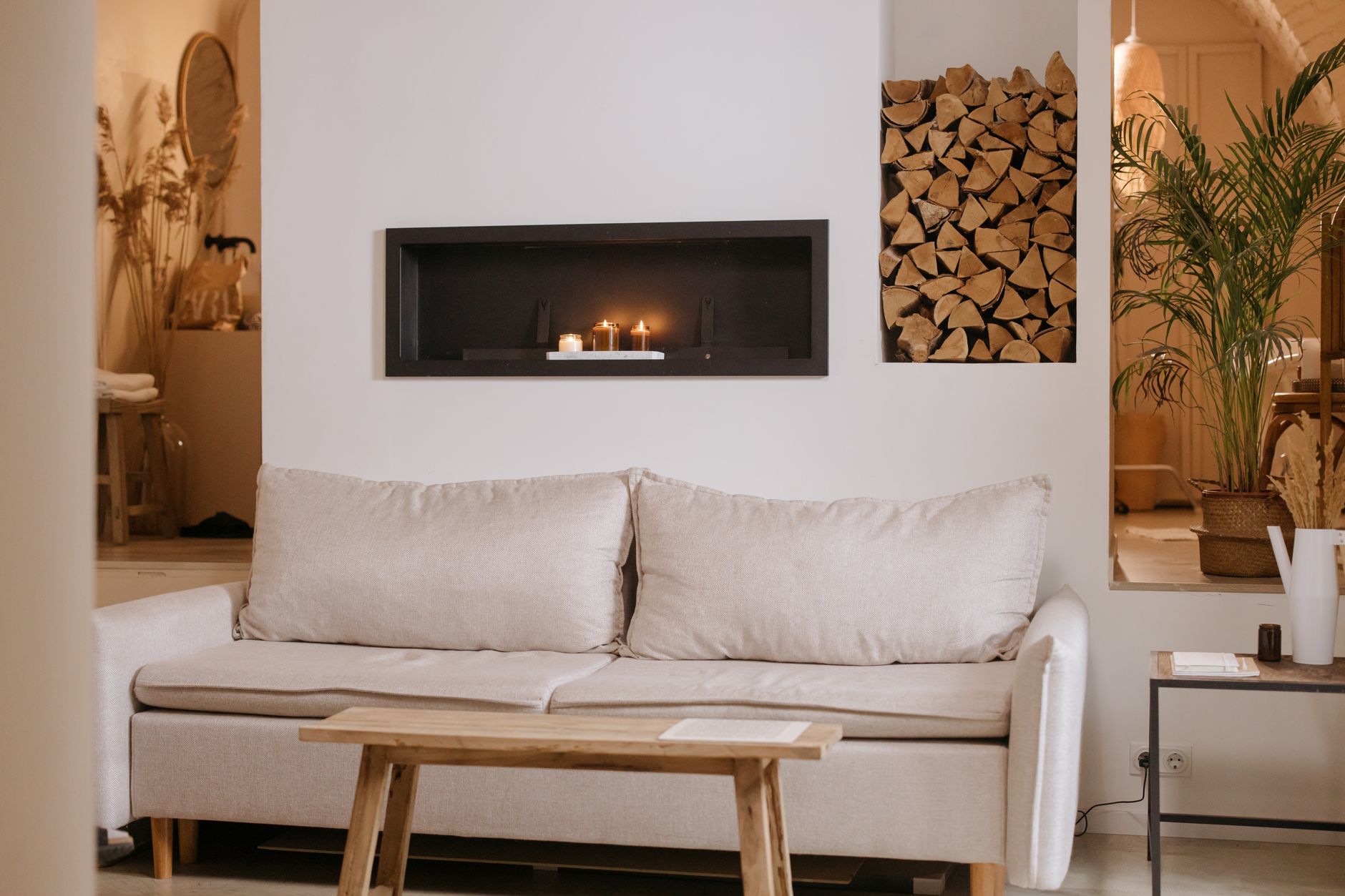 Salon minimaliste aux tons neutres avec canapé beige, table basse en bois, cheminée encastrée avec bougies et niche de bûches.